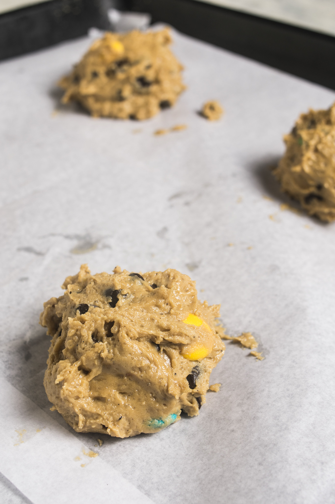 Portions of browned butter chocolate chip and M&M cookie dough arranged on a lined baking tray.